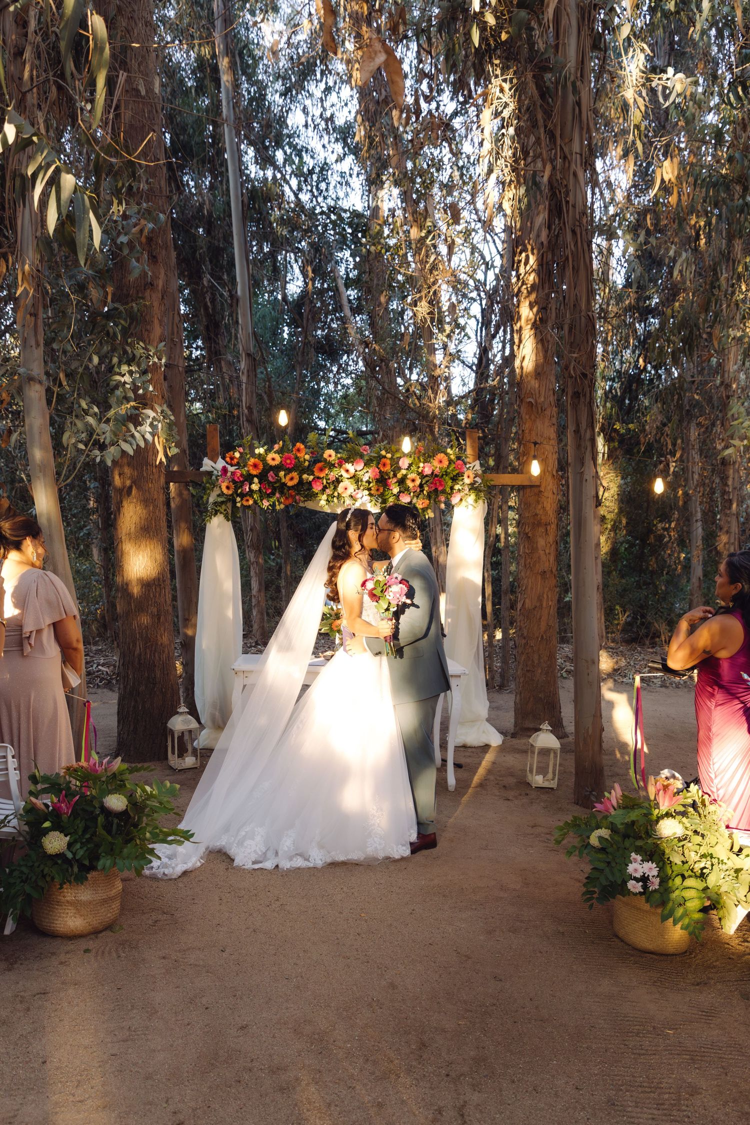 Bride and groom share first kiss under floral arch surrounded by tall trees at forest wedding.
