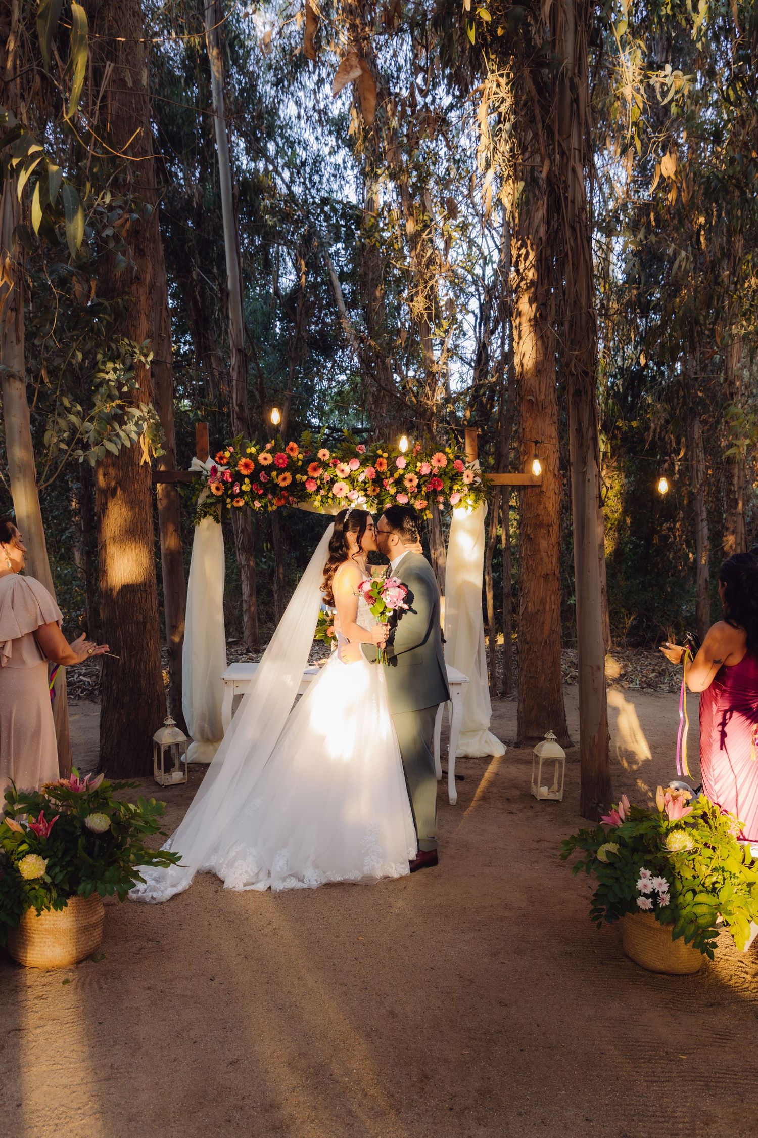 Newlyweds kissing under decorated floral arch with string lights in enchanting forest wedding venue.