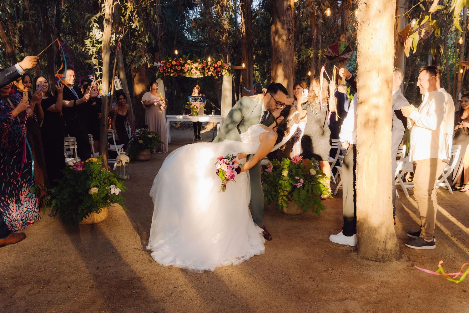 Groom dipping bride for romantic kiss while guests celebrate at outdoor woodland wedding reception.