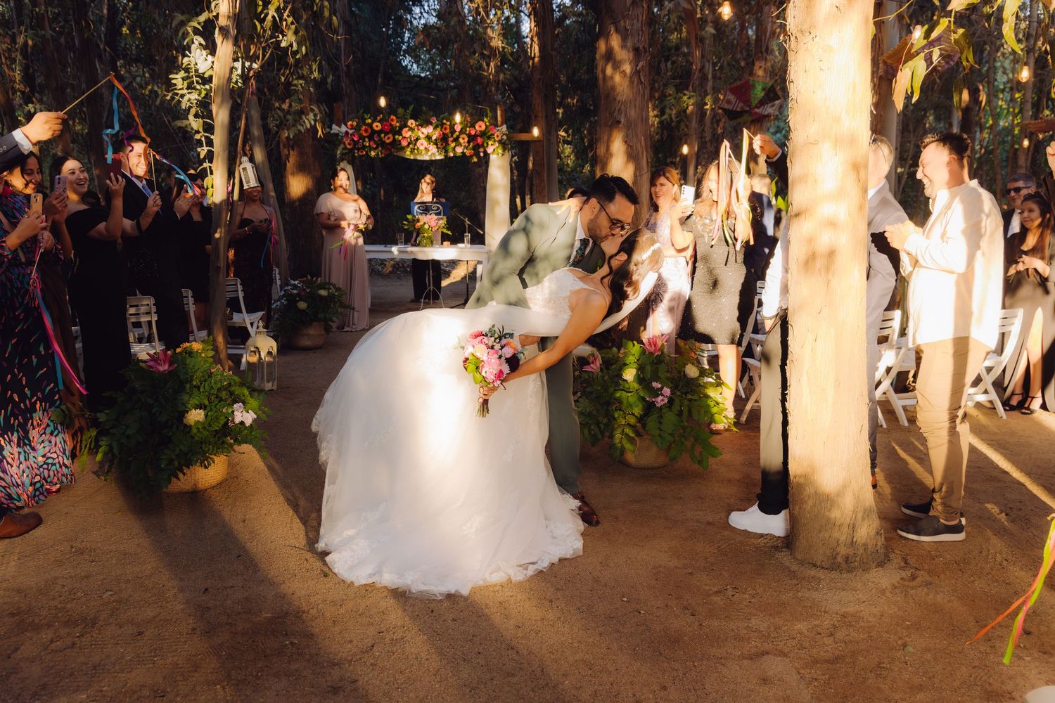 Newlyweds sharing romantic dip kiss surrounded by cheering guests at elegant outdoor forest wedding.
