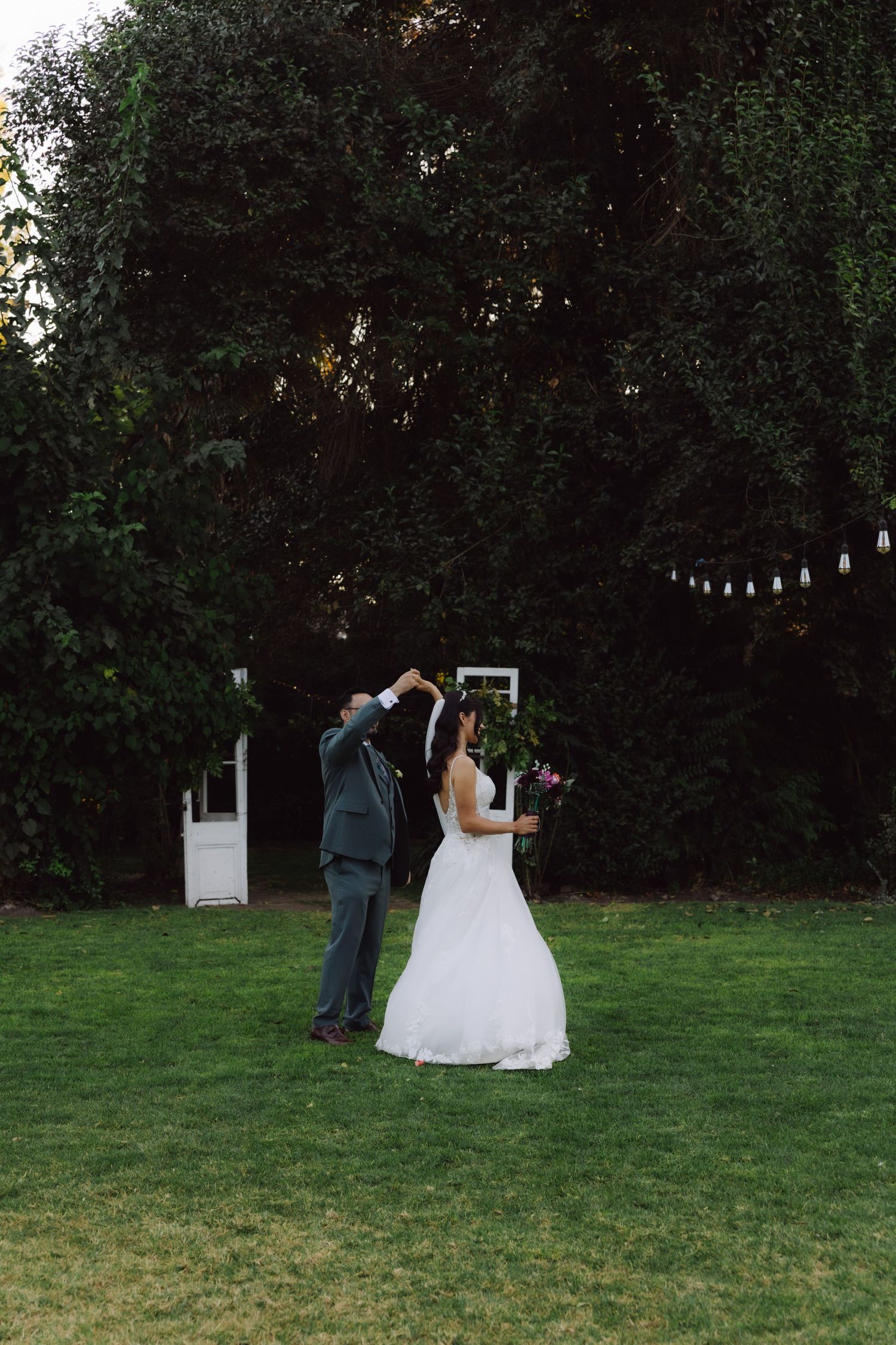 Newlywed couple dancing on lush green lawn with vintage white doors backdrop at garden wedding.