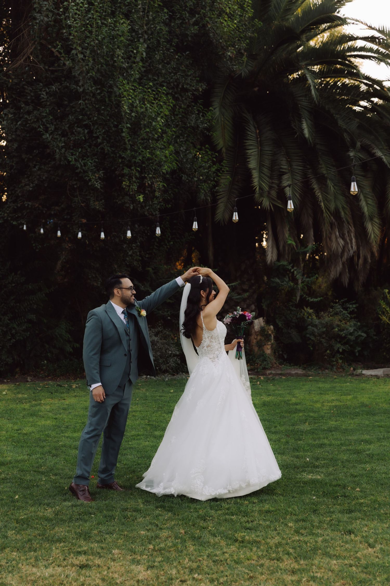 Groom twirling bride in white wedding gown on lush green lawn with string lights and palm trees.