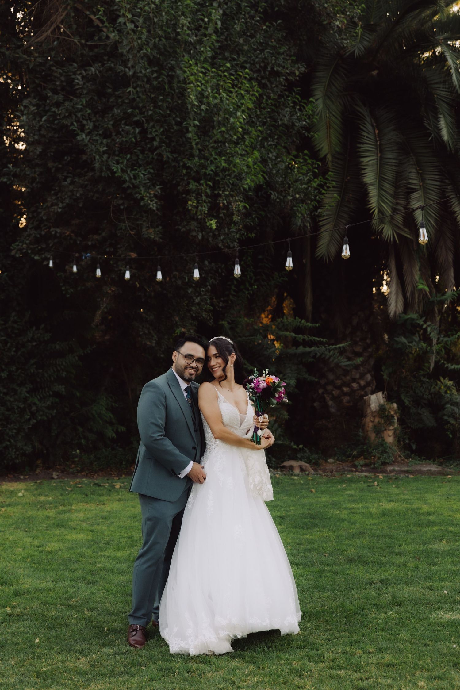 Newlyweds posing together on green lawn with colorful bridal bouquet and string lights backdrop.