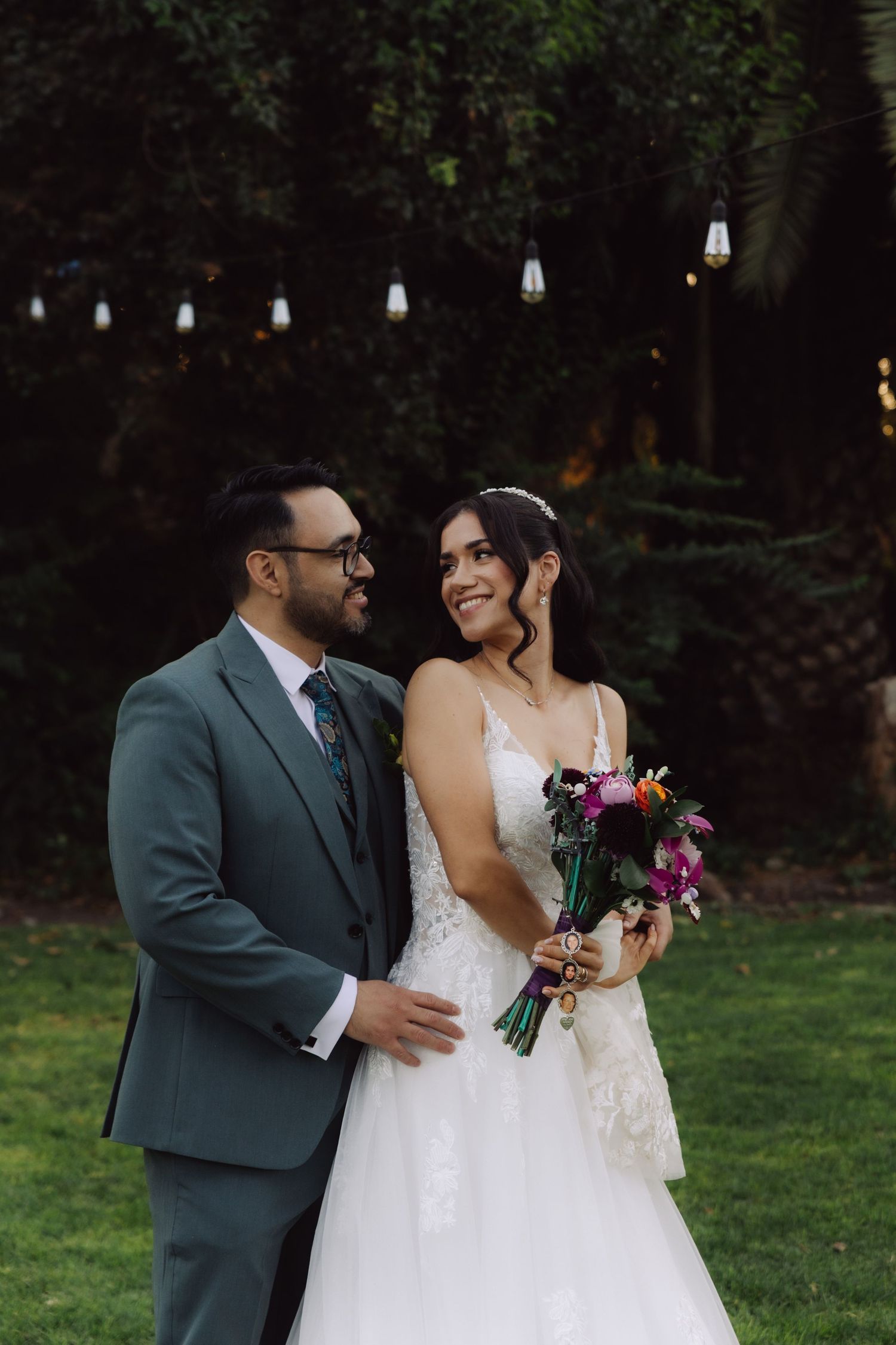 Bride and groom sharing a romantic glance outdoors with colorful bouquet and string lights.