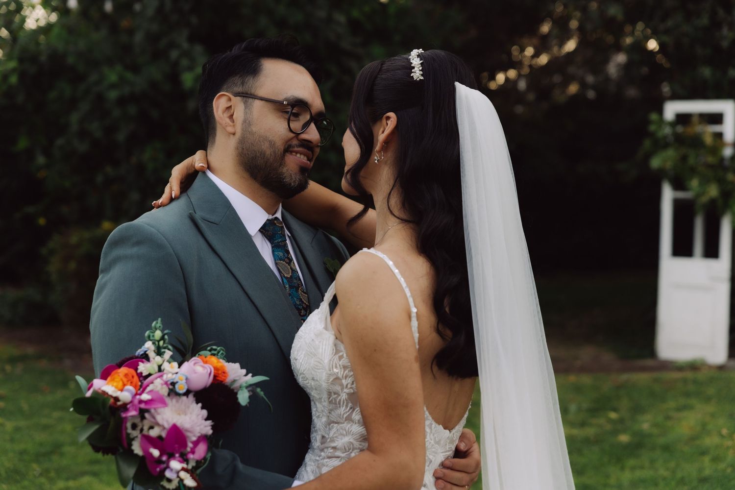 Bride and groom in intimate embrace with colorful floral bouquet at outdoor wedding venue.