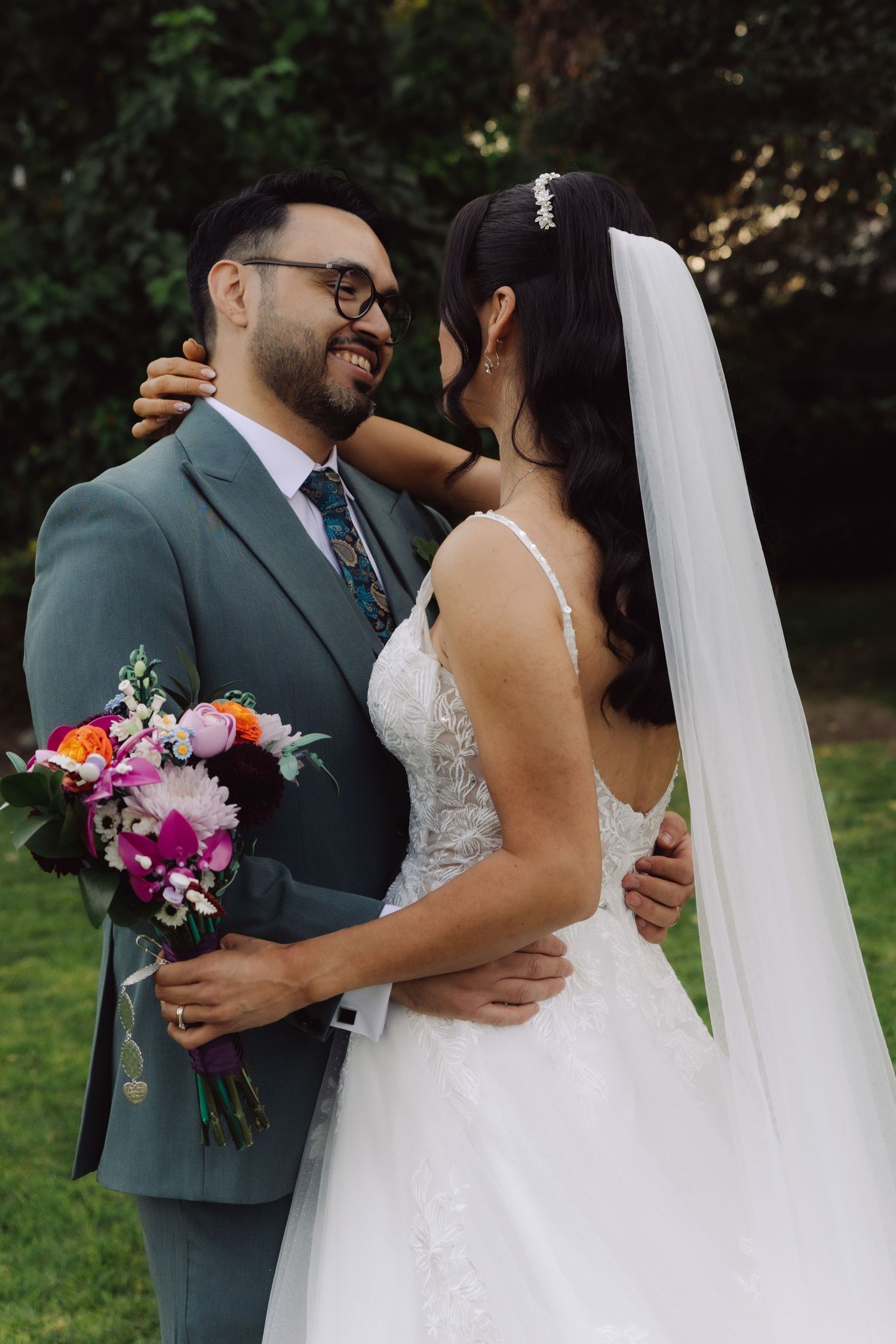 Smiling groom holds colorful bouquet while embracing bride in white gown with veil outdoors.