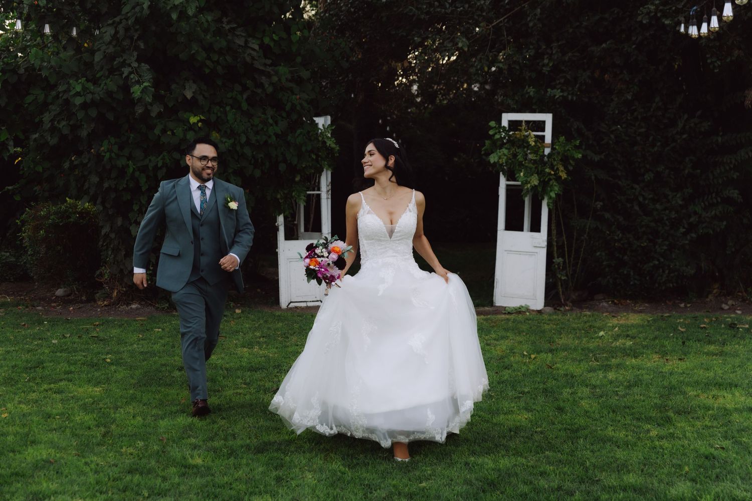 Bride in white gown and groom in gray suit walking on green lawn at outdoor wedding.