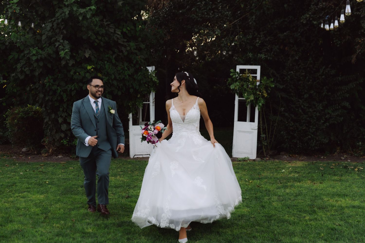 Bride in white ball gown walking on green lawn with groom in gray suit at garden wedding.