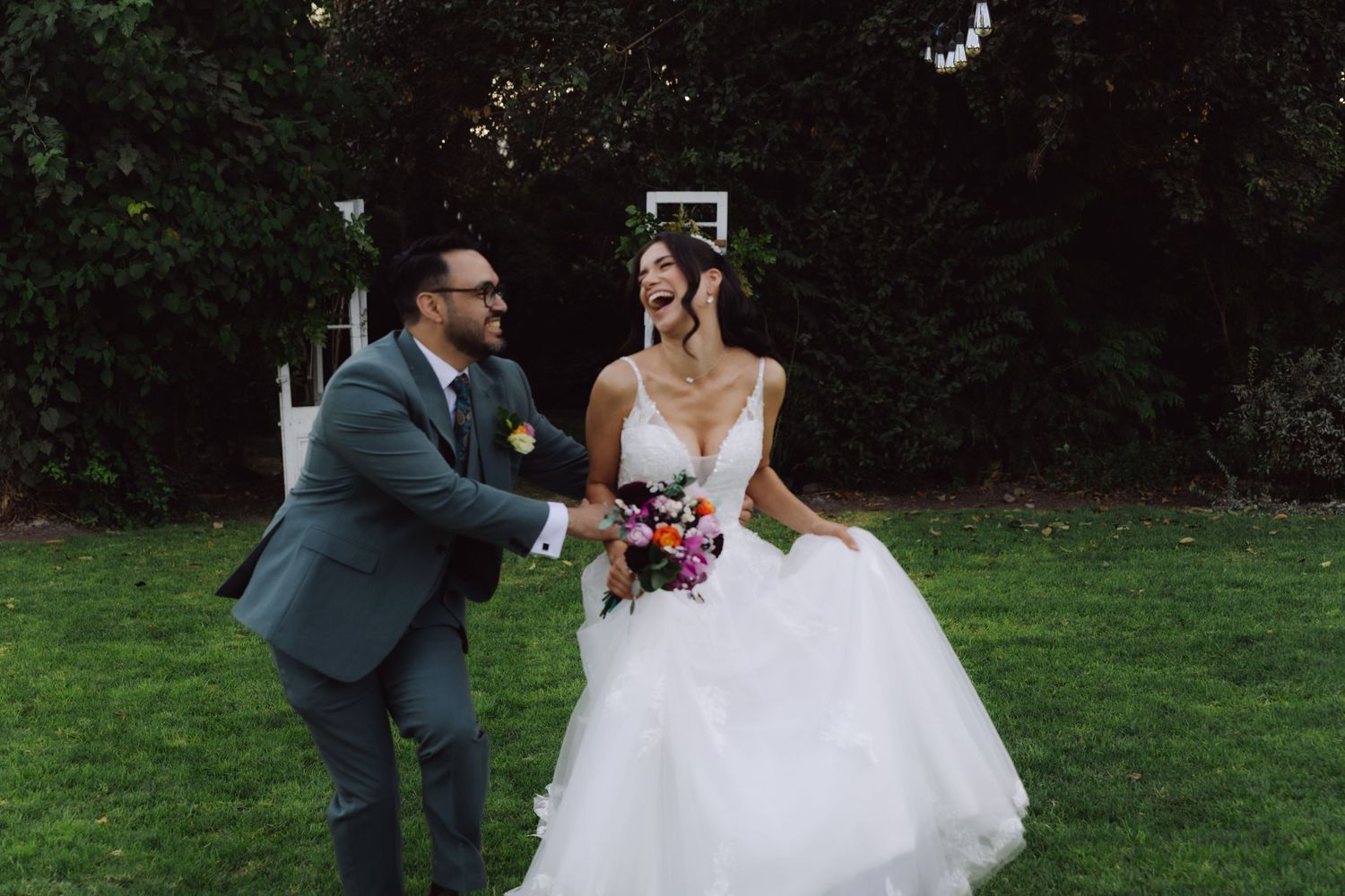 Laughing bride and groom sharing joyful moment on green lawn during outdoor wedding celebration.