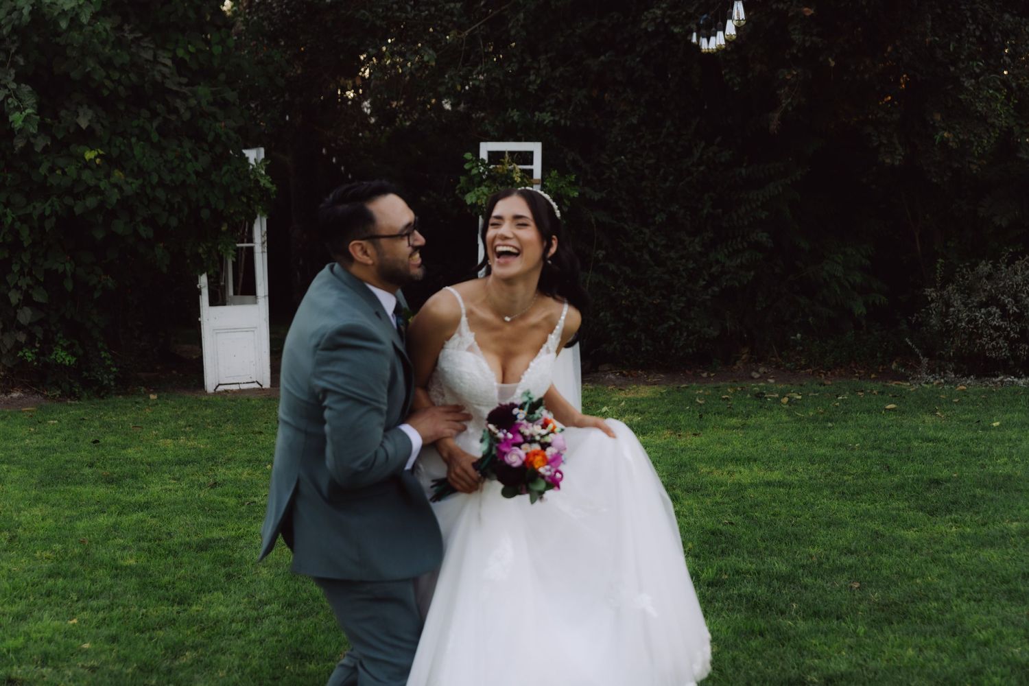 Bride laughing joyfully with groom holding colorful bouquet during outdoor wedding garden session.