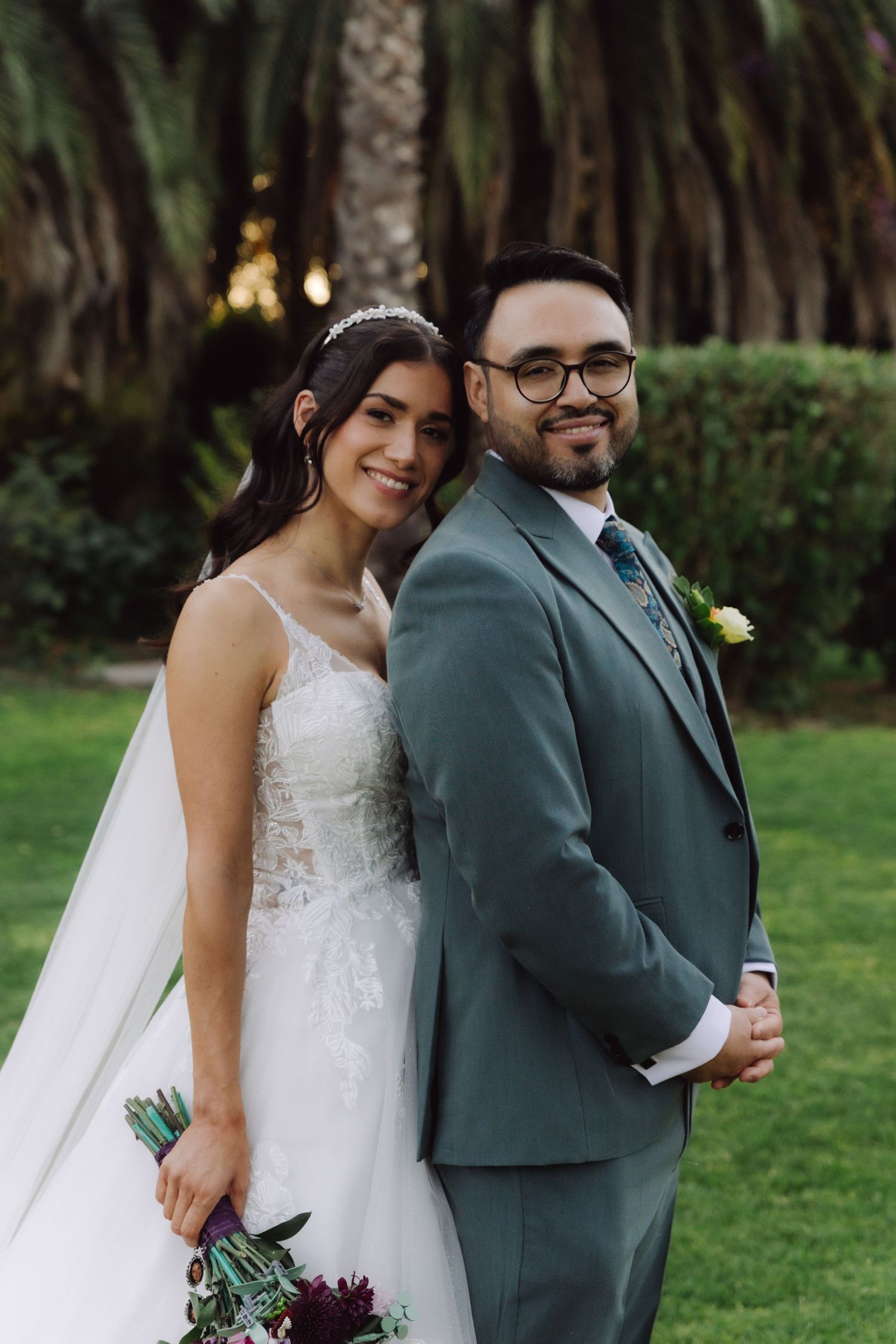Bride and groom posing back to back surrounded by palm trees at outdoor garden wedding.