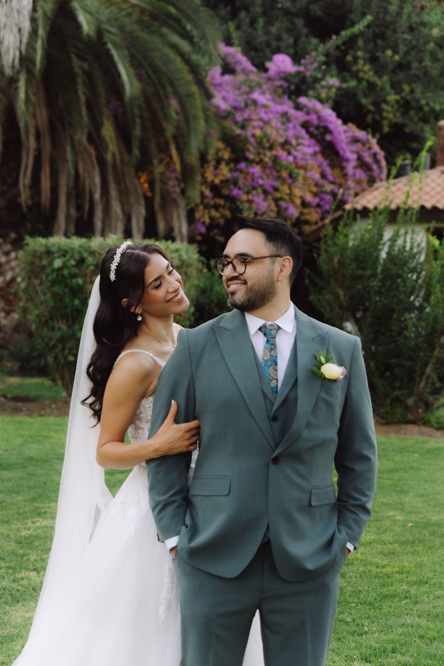 Bride and groom standing together in garden with purple bougainvillea blooms in background.