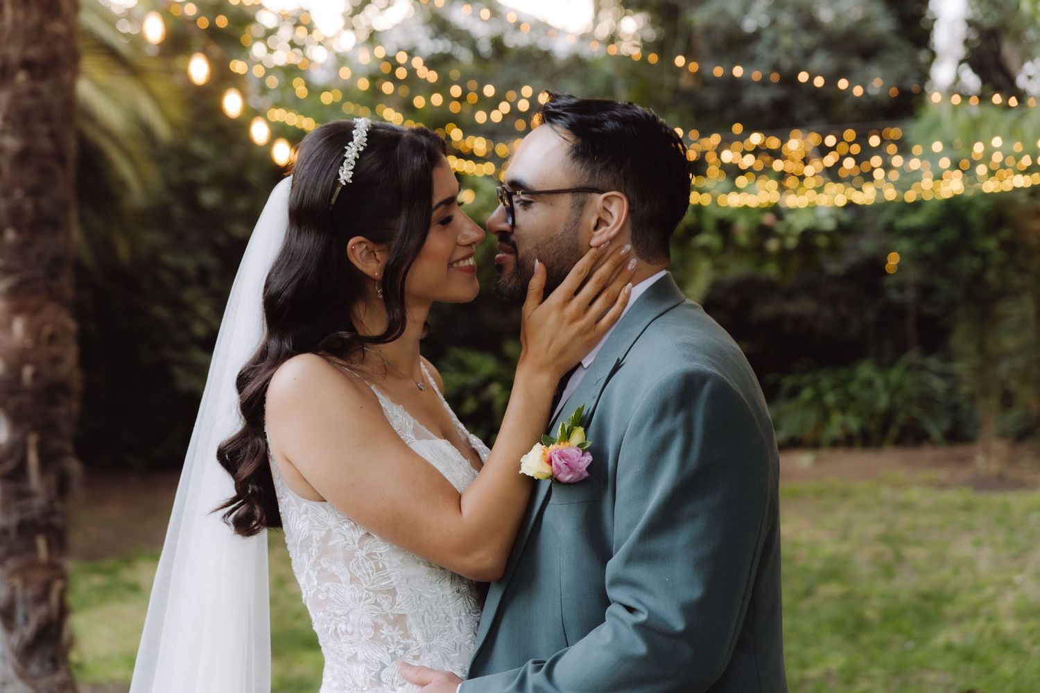 Bride and groom nearly kissing under warm string lights surrounded by greenery at their wedding.