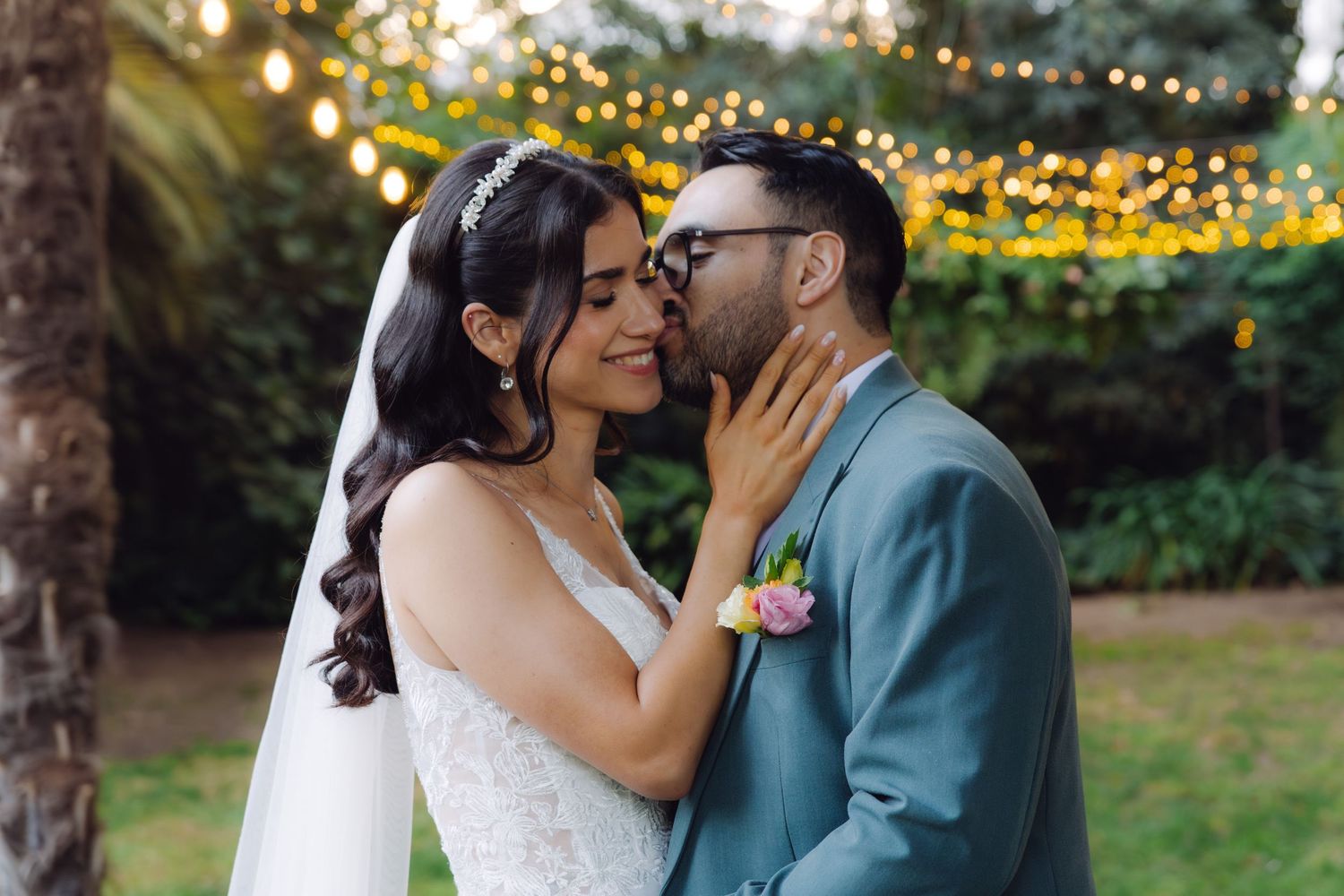 Smiling bride and groom in teal suit sharing an intimate moment under bokeh string lights outdoors.