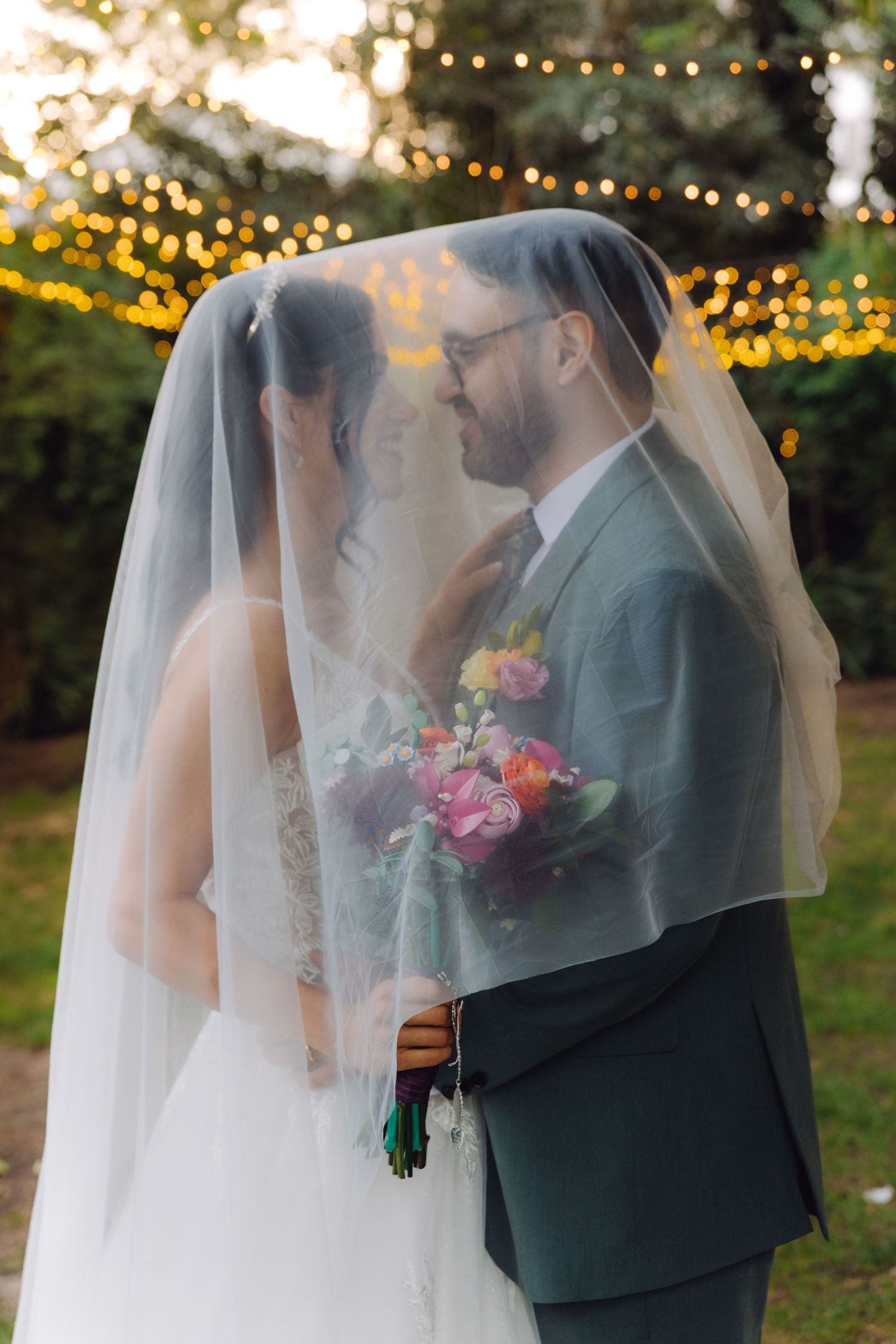 Bride and groom standing close together under a flowing white veil with colorful bouquet at garden wedding.