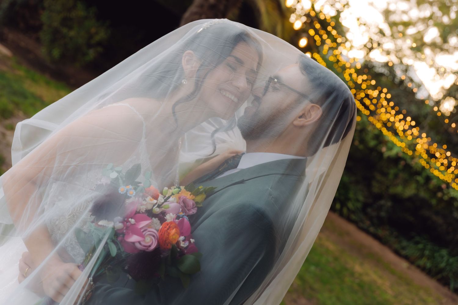 Bride and groom smiling under a sheer veil holding a vibrant pink and orange bouquet at sunset.