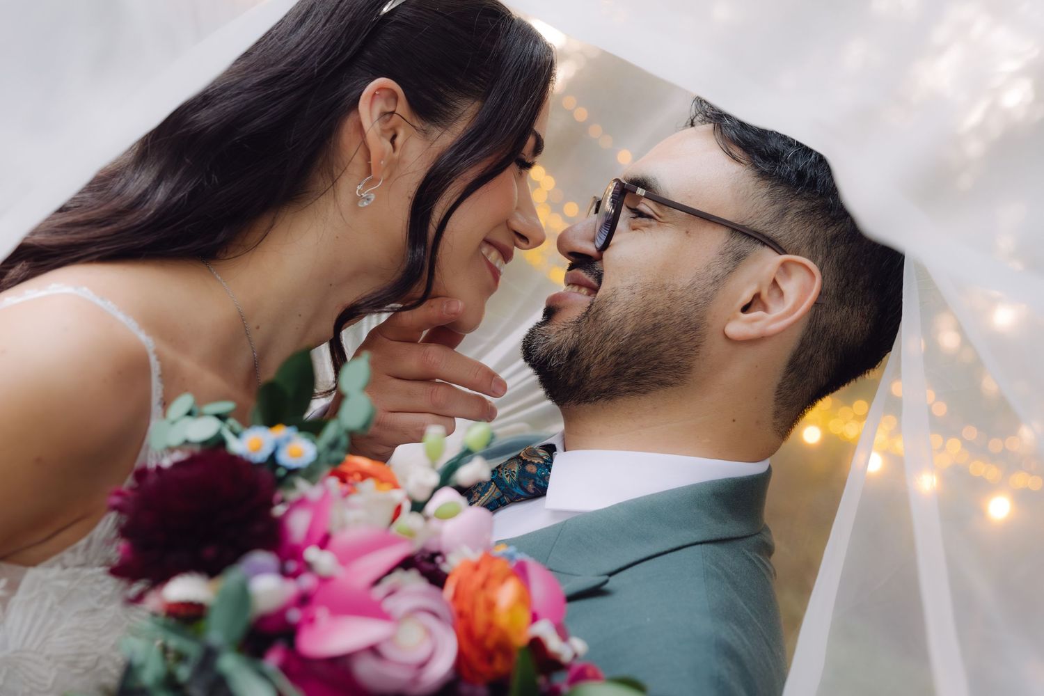 Bride and groom sharing an intimate close-up moment under a veil with a colorful floral bouquet.