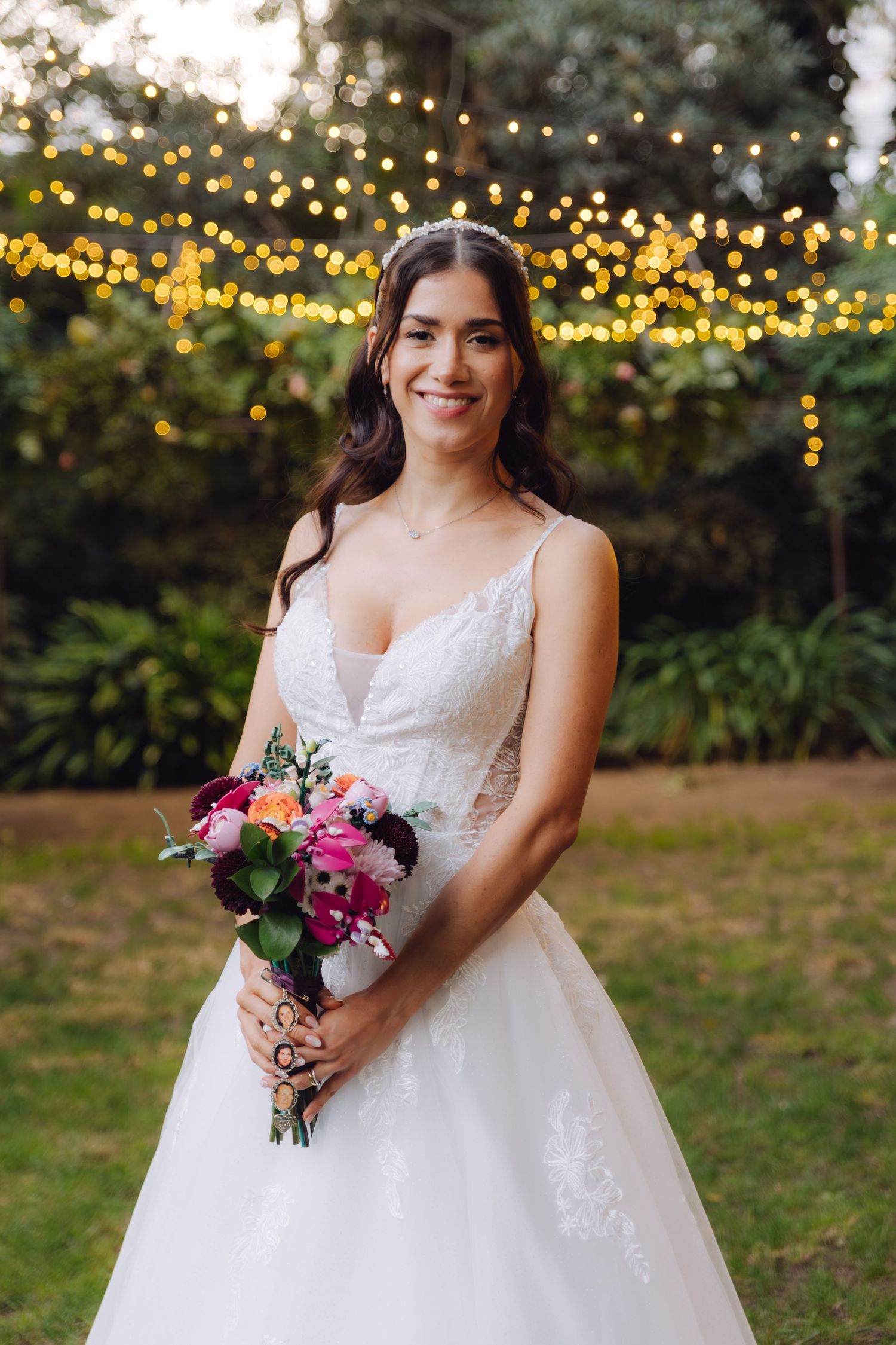 Smiling bride in lace wedding gown holding a vibrant pink and purple bouquet under garden string lights.