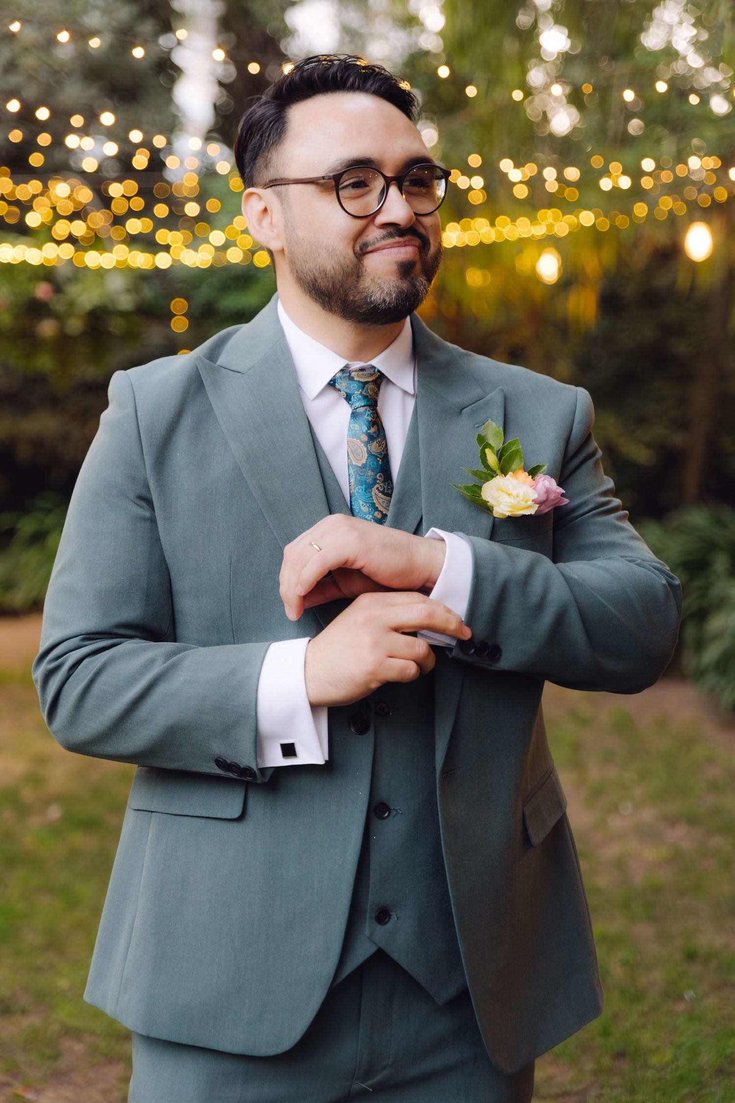 Dapper groom in teal three-piece suit adjusting cufflinks with floral boutonniere under string lights.