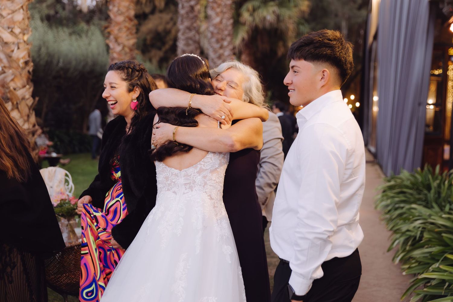 Bride in white wedding gown receives warm hugs from smiling guests at an outdoor wedding reception.