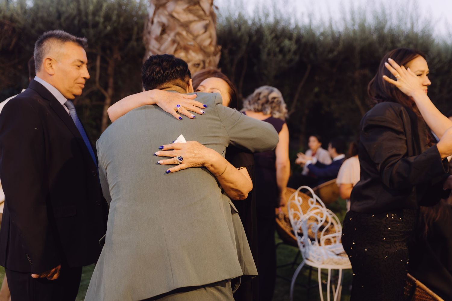 Two men share an emotional embrace at an outdoor wedding ceremony while guests mingle nearby.