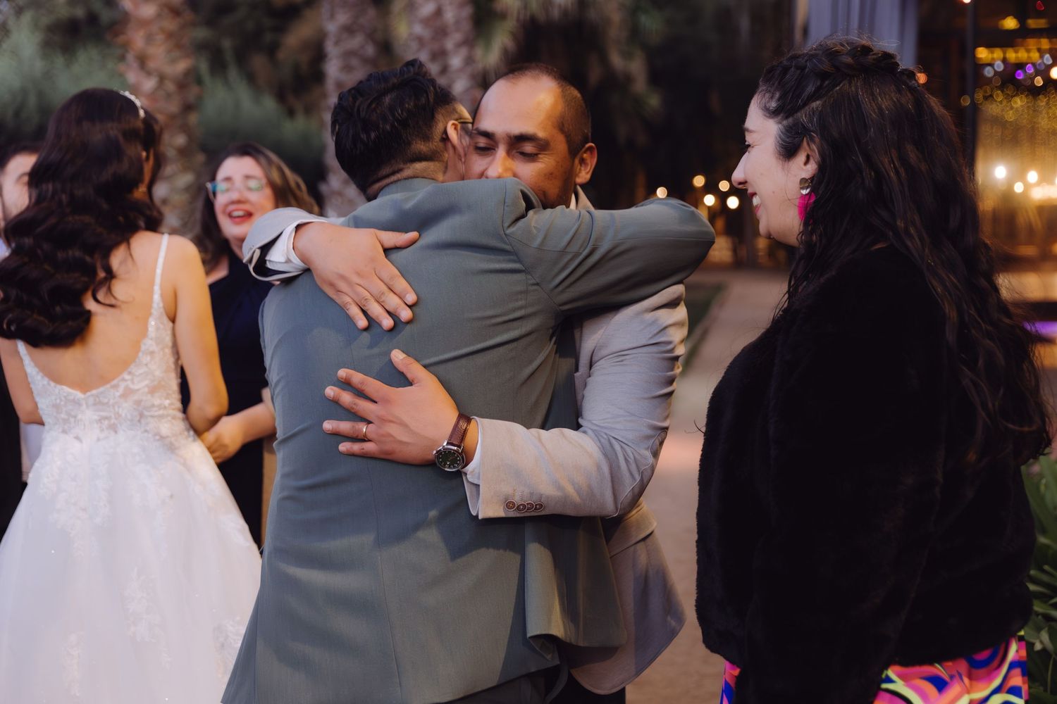 Groom in gray suit shares a heartfelt hug with a guest at an outdoor wedding reception.