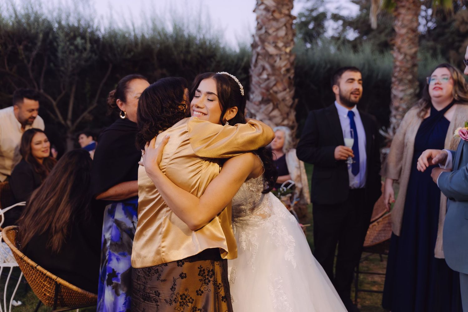 Smiling bride in white gown embraces a guest in gold dress at an outdoor wedding celebration.