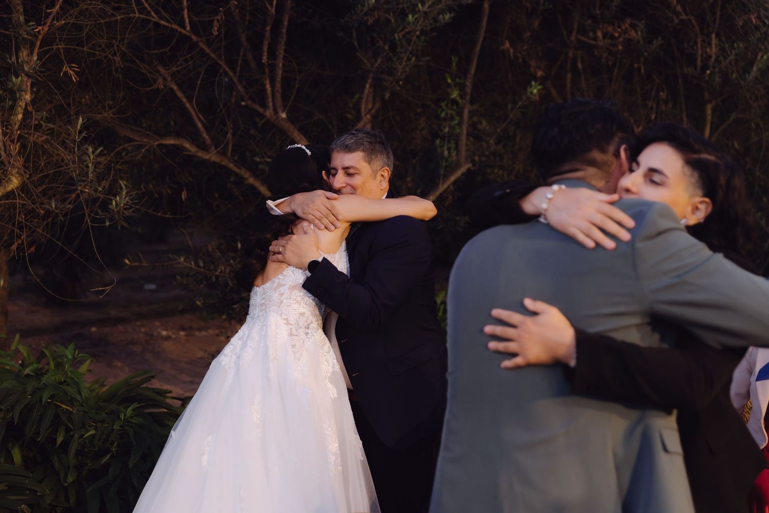 Bride and groom share simultaneous hugs with guests in a touching outdoor wedding moment.