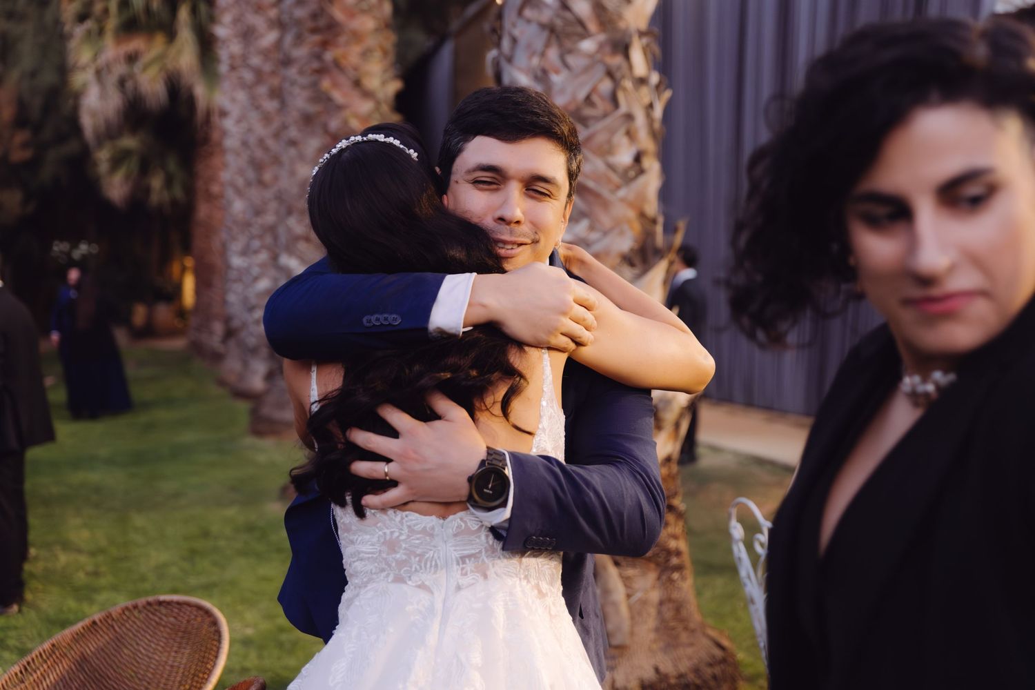 Groom in blue suit smiles while embracing a guest warmly at an outdoor wedding reception.