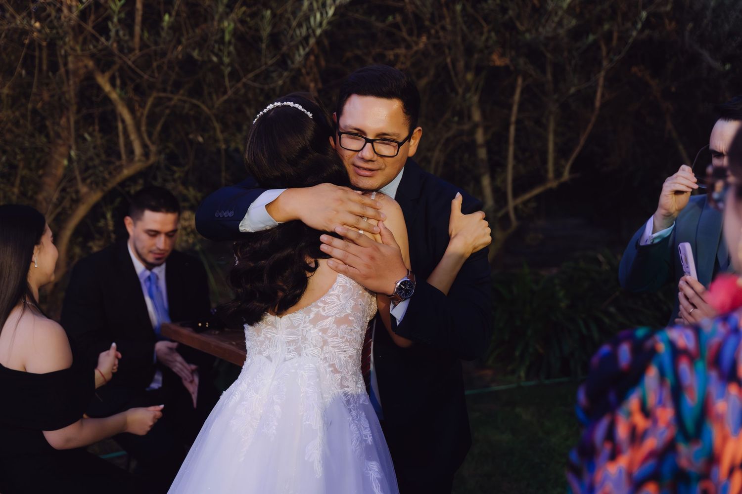 Bride in white lace gown shares a warm hug with a bespectacled guest at a wedding.