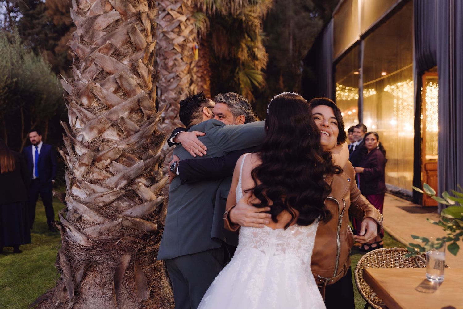 Bride in white wedding dress smiles as groom hugs a guest near a palm tree outdoors.
