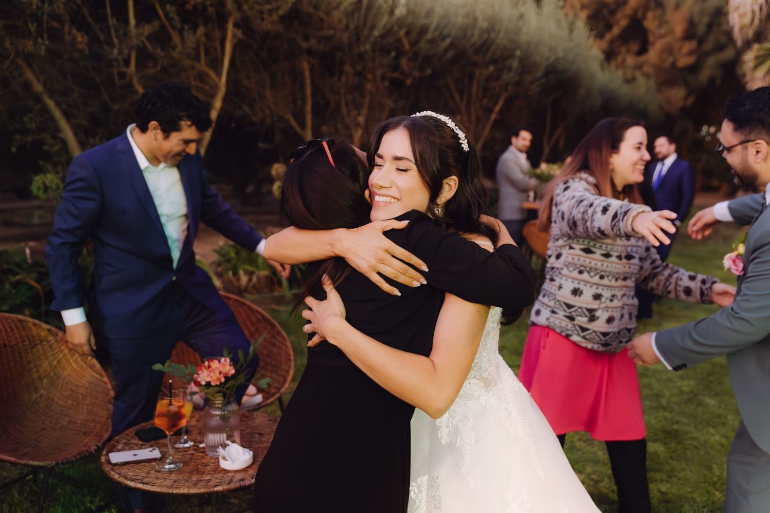 Beaming bride in white gown embraces a guest in black at an outdoor wedding reception.