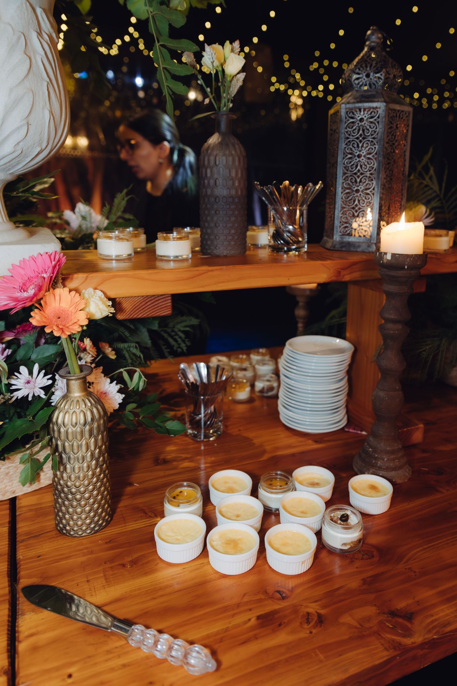 Wedding dessert table with creme brulee ramekins, stacked plates, candles, and floral arrangements at night.