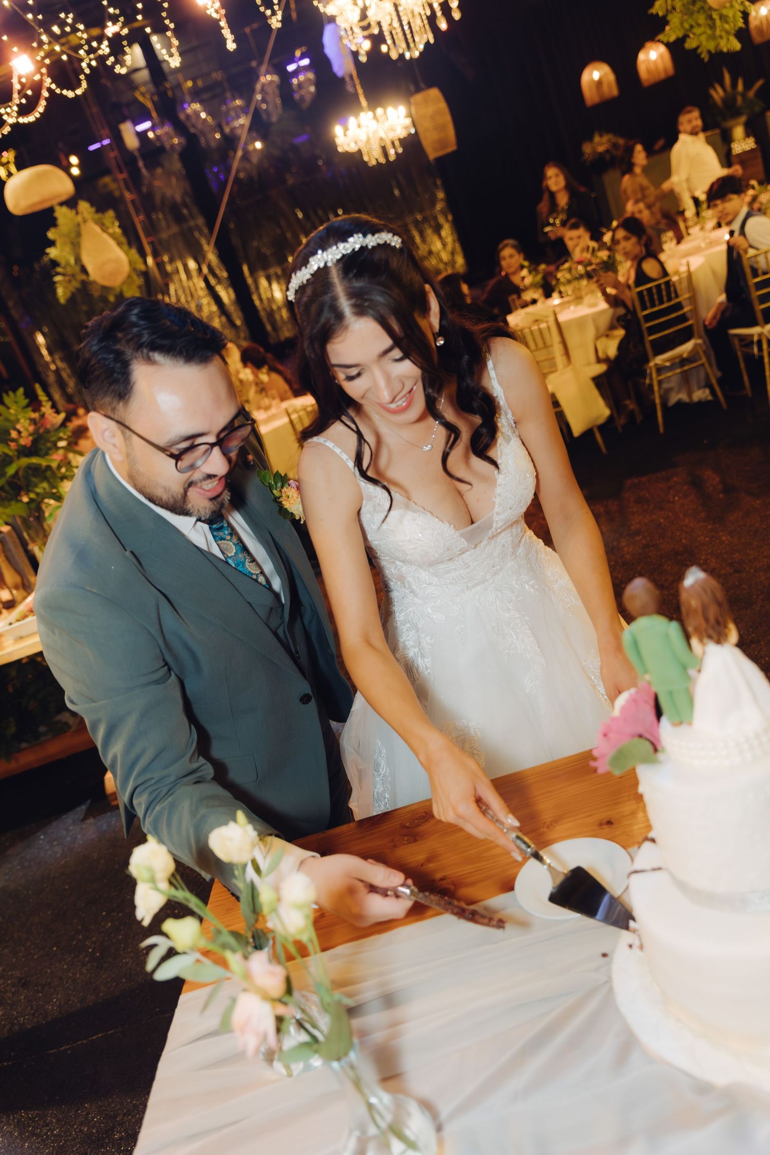 Smiling bride and groom cutting their white wedding cake together at an outdoor evening reception.