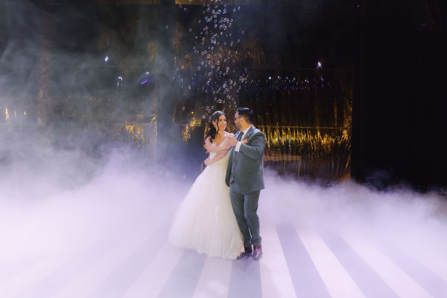 Bride in white ballgown and groom dance romantically in thick fog under sparkling lights at wedding reception.