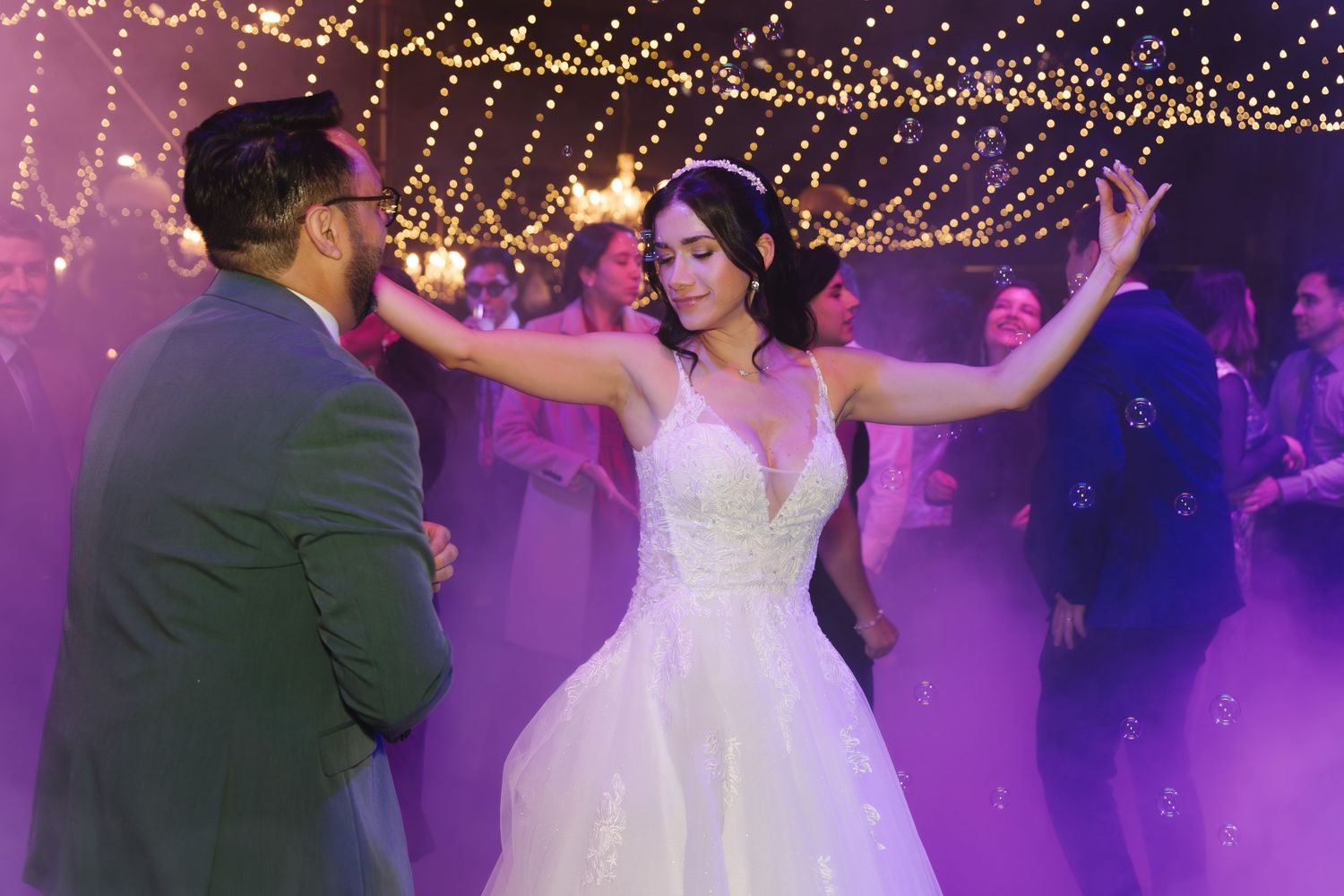 Bride in stunning white gown dances joyfully in purple fog surrounded by bubbles at wedding reception.