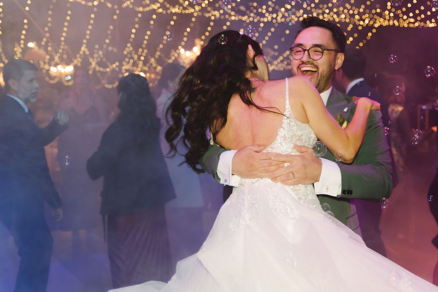 Laughing bride and groom share joyful embrace while dancing under string lights at festive wedding reception.