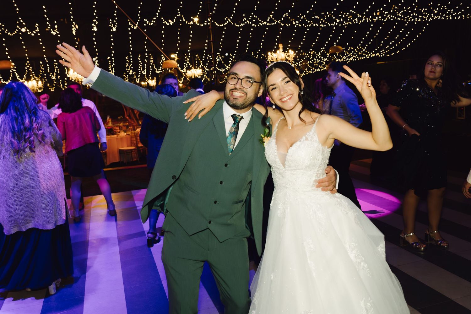 Cheerful bride and groom pose playfully on dance floor under string lights at lively wedding reception.