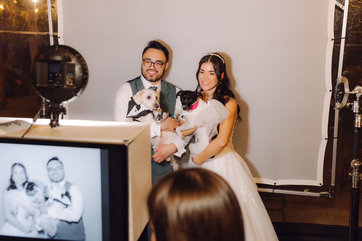 Bride and groom smile holding small dogs during fun wedding photo booth session with monitor preview.