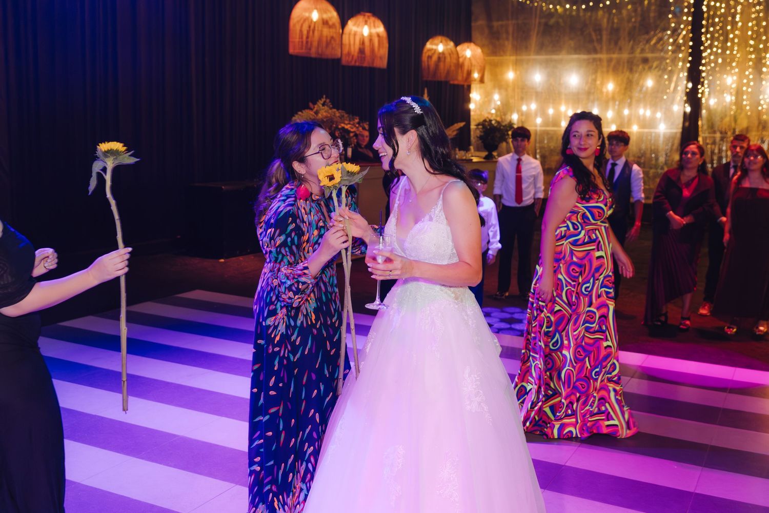 Bride in white gown shares a toast and conversation with colorfully dressed guest on wedding dance floor.