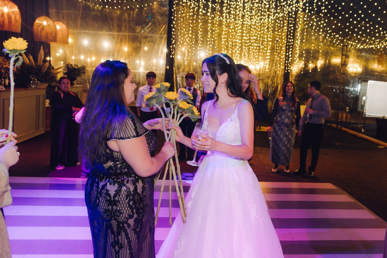 Bride in white gown chatting with guest on dance floor under warm string lights at reception.