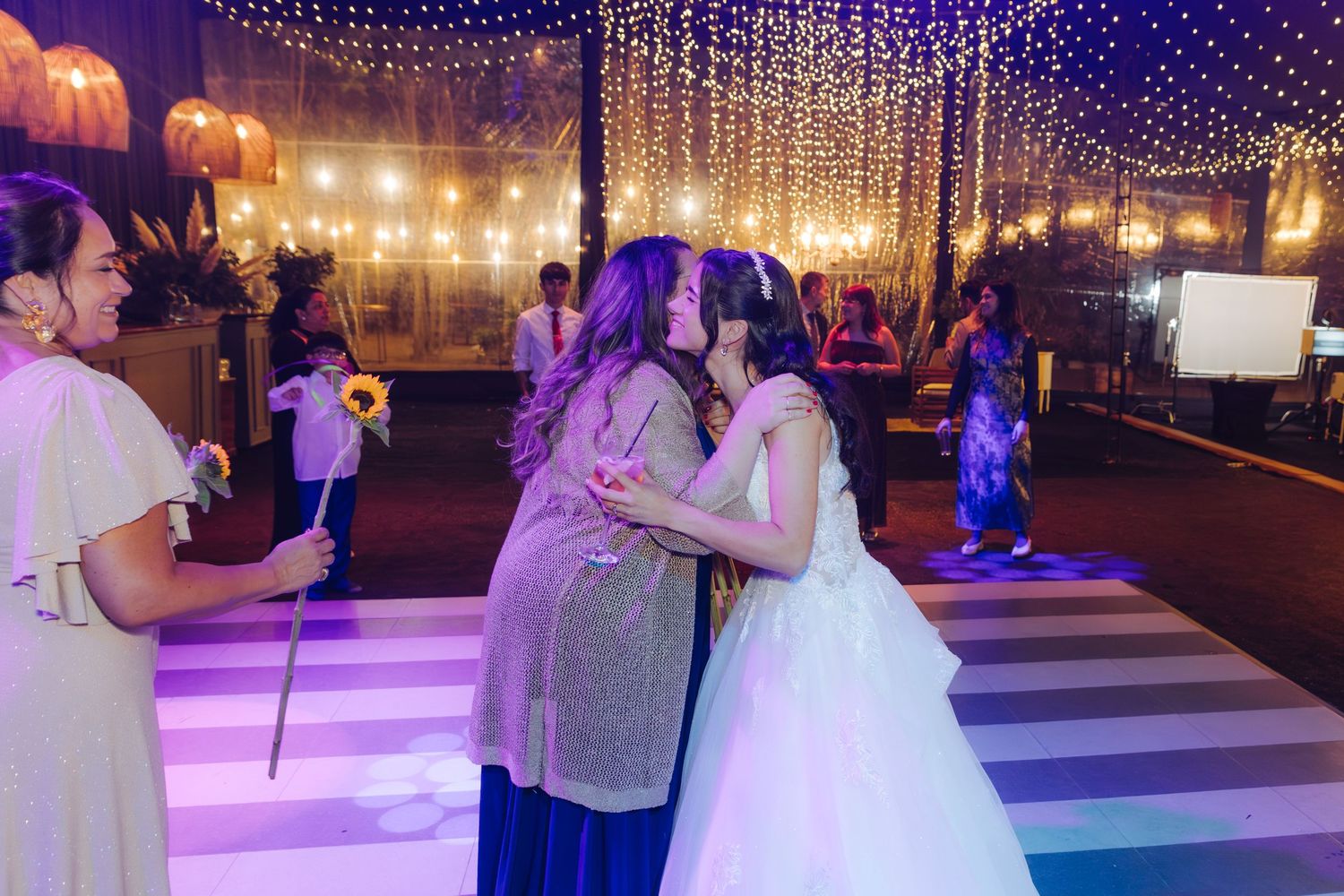 Bride shares a warm kiss with pregnant guest on striped dance floor at joyful wedding reception.