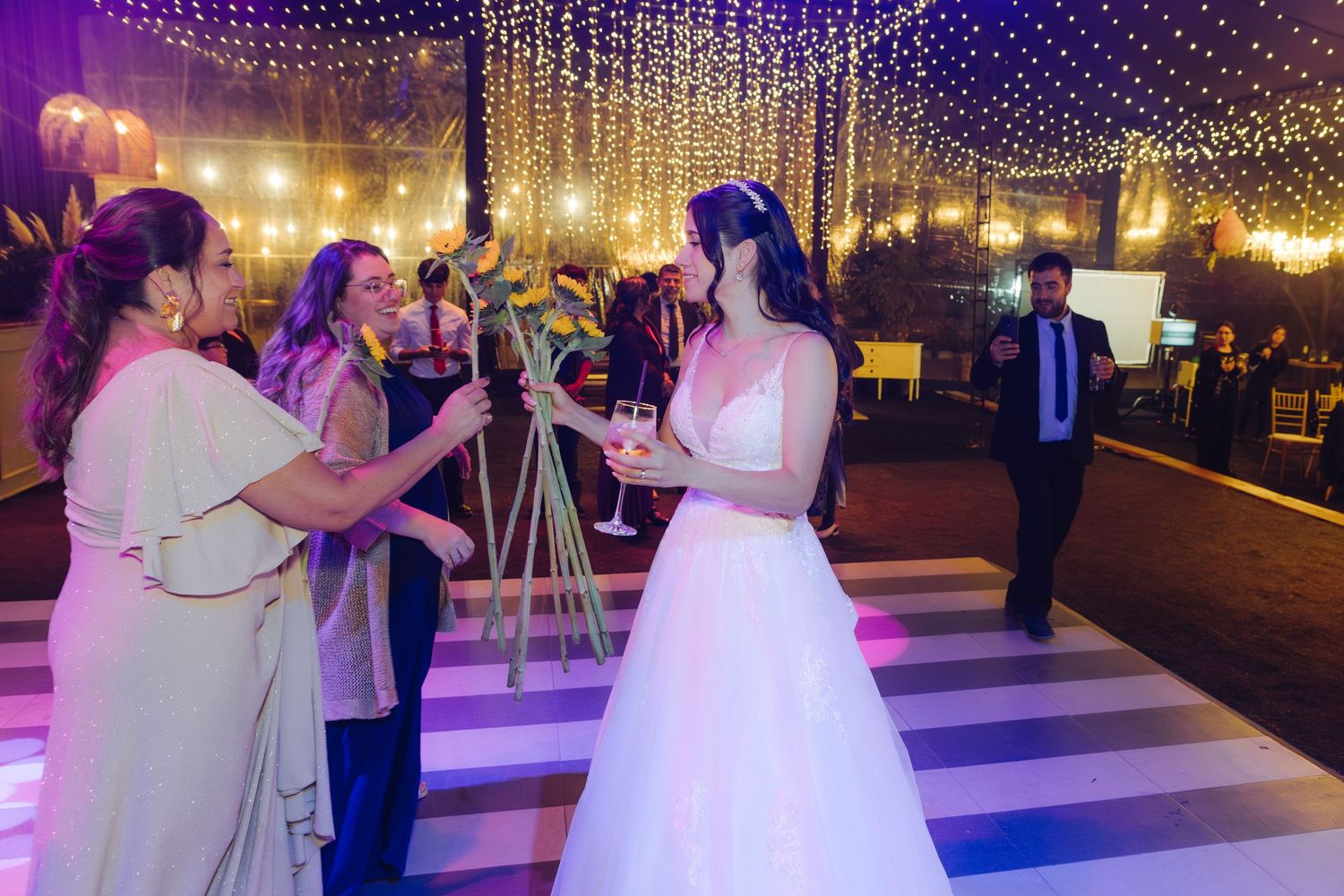 Bride in white gown toasting with guests on striped dance floor under fairy lights at elegant wedding reception.