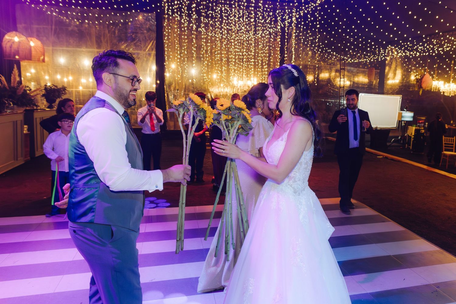 Bride and groom dancing together on striped dance floor under sparkling string lights at reception.
