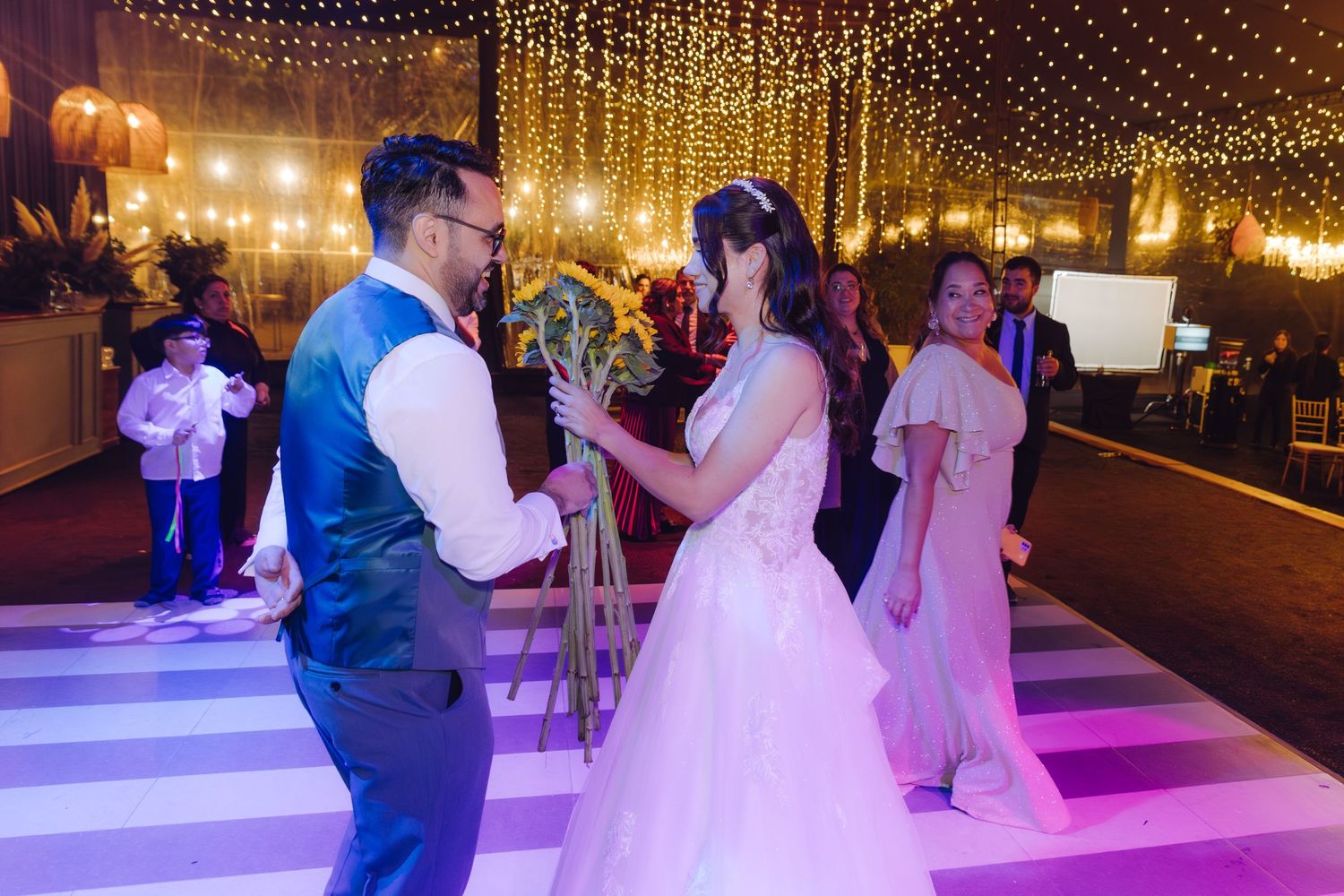 Groom in blue vest and bride in white gown share first dance under sparkling fairy lights at wedding reception.