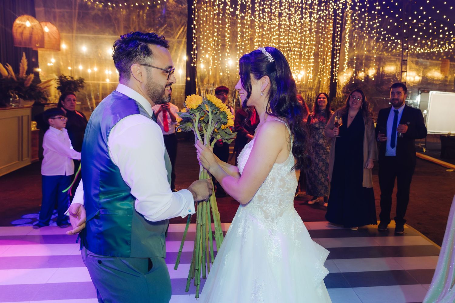 Bride and groom share romantic first dance moment with colorful sunflower bouquet under twinkling wedding lights.