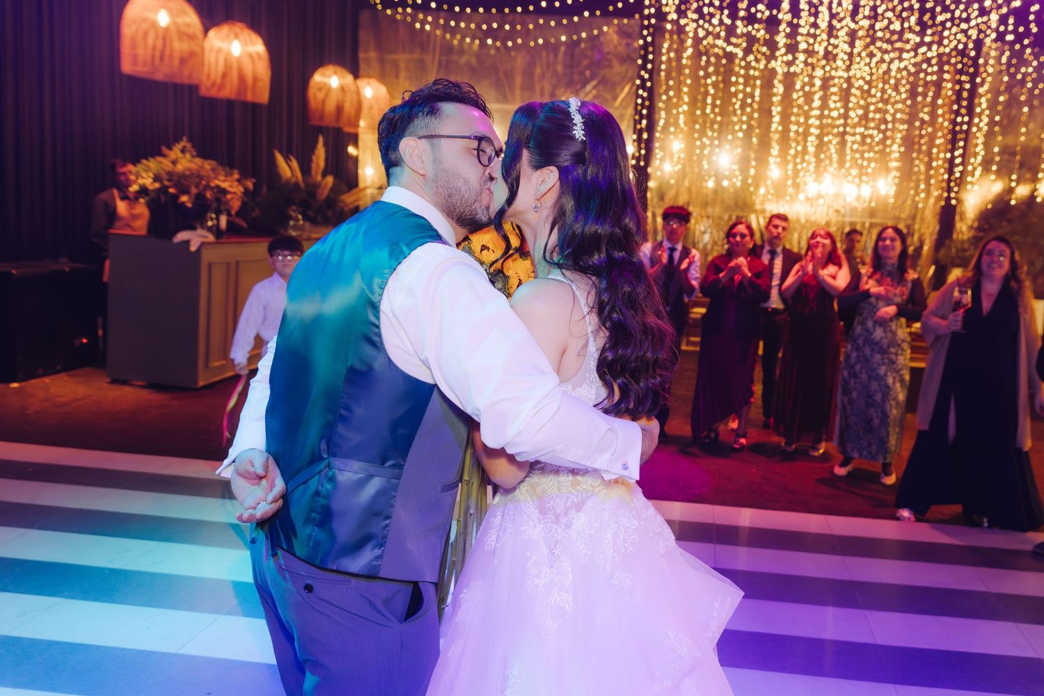 Newlyweds sharing first kiss on dance floor surrounded by guests under romantic string lights.