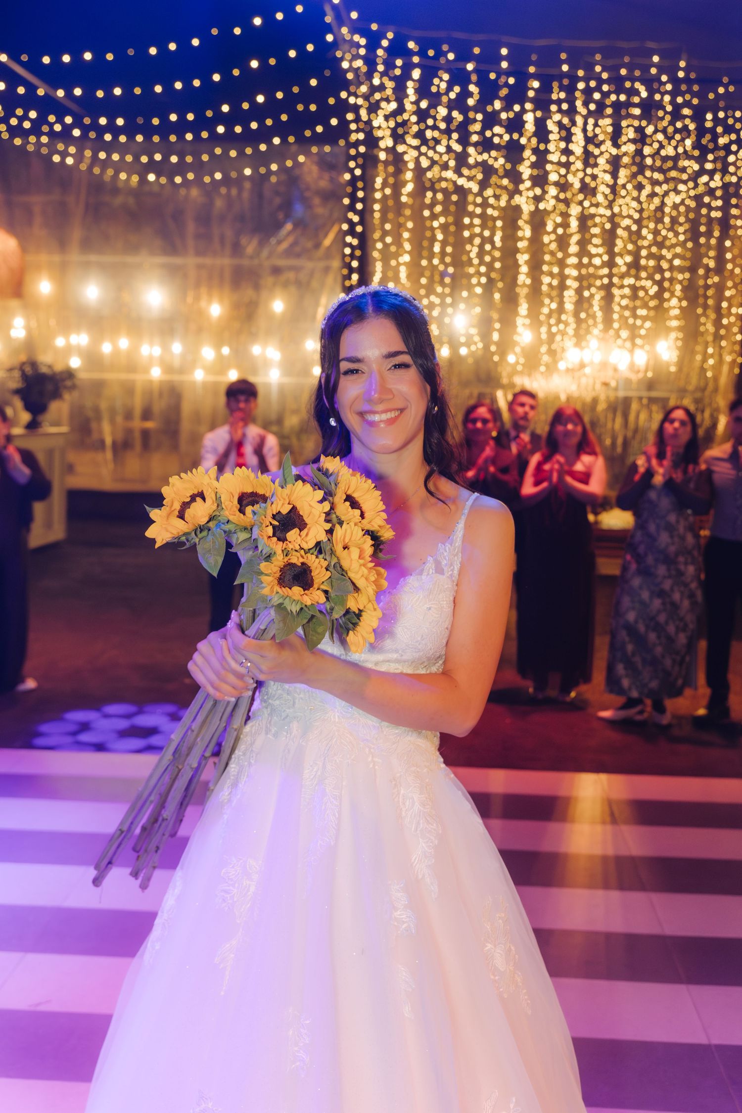 Smiling bride in white lace gown holding sunflower bouquet on dance floor with fairy lights backdrop.