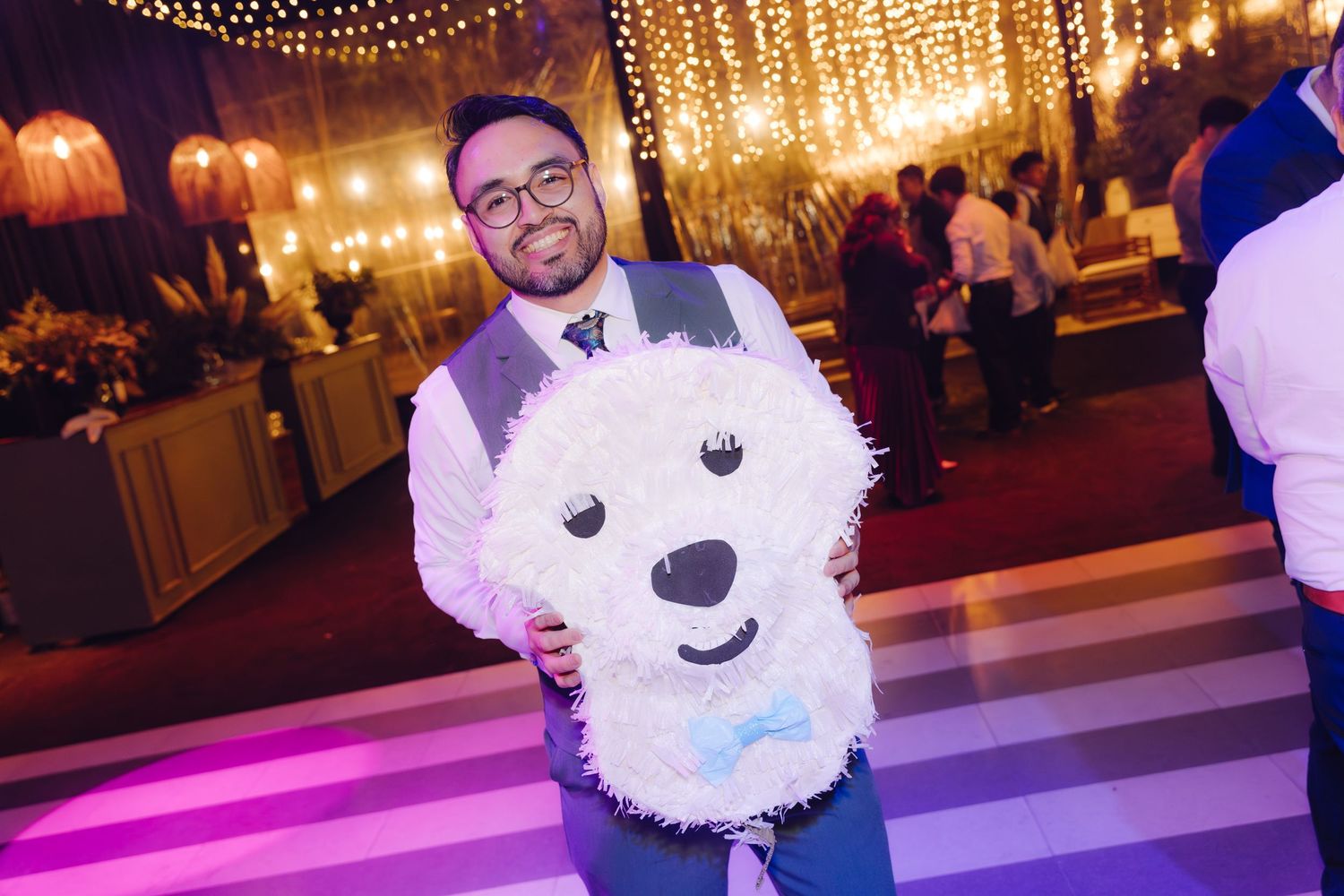 Smiling man in suit holding white fluffy bear-shaped pinata on decorated wedding dance floor.