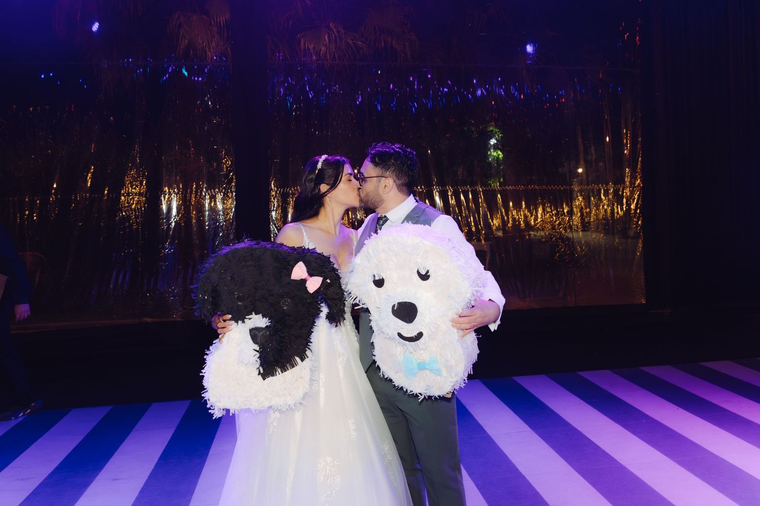 Bride and groom sharing a kiss while holding decorative dog pinatas on a striped dance floor.